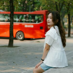 A young woman sits on a pavement bench in the park with a red bus in the background.